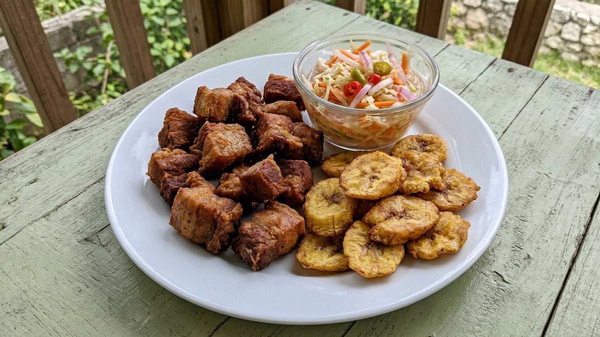 A plate of Haitian food on a home table: golden fried griot pork, fried plantain and a bowl of bright pikliz cabbage slaw