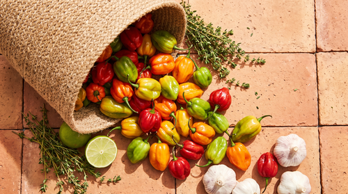 Ripe scotch bonnet chillies spilling from a woven basket onto terracotta, ranging from green through yellow and orange to deep red, with thyme, lime and garlic